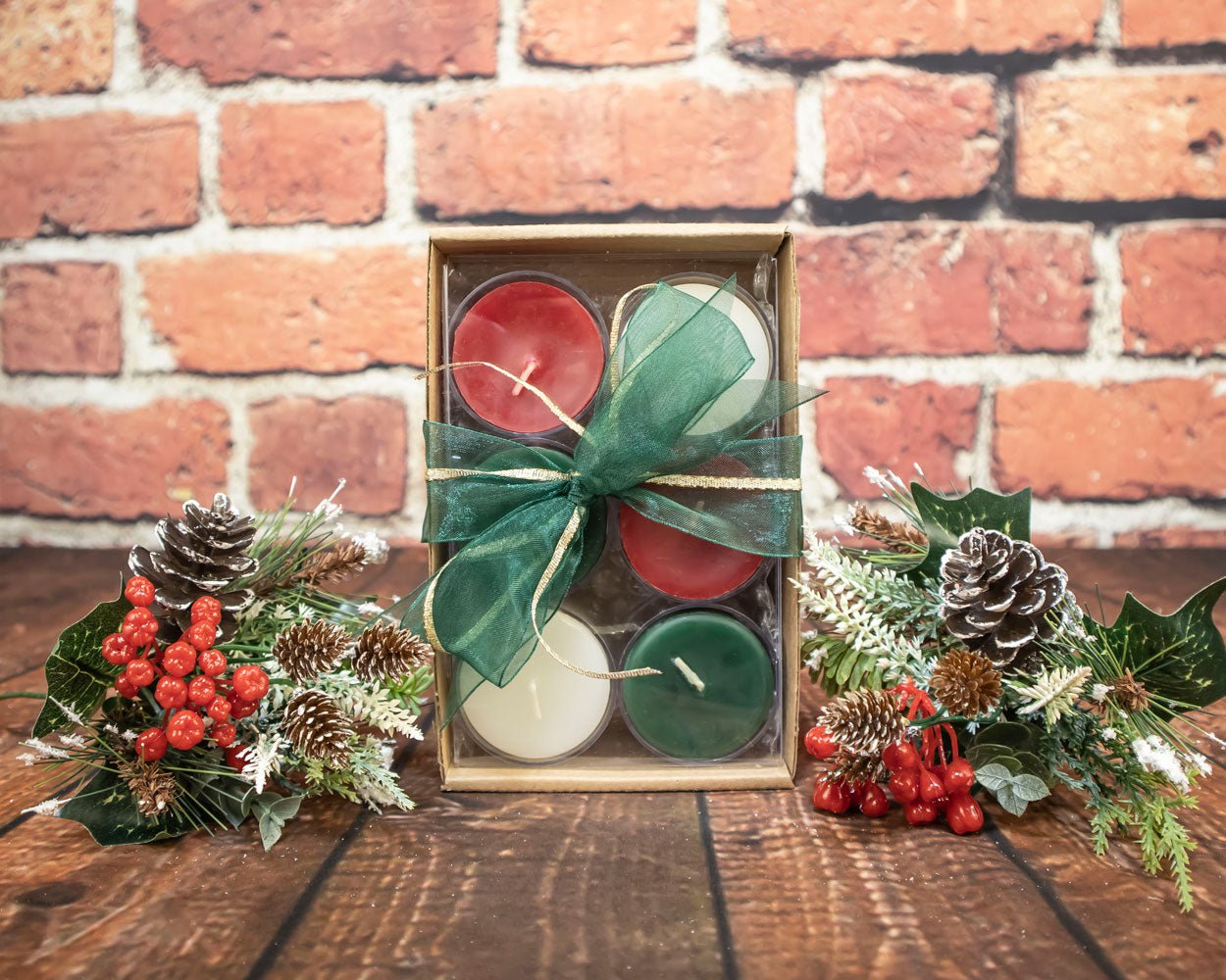 Set of four candles in a decorative box with green ribbon, surrounded by festive decorations on a wooden surface with a brick wall background.